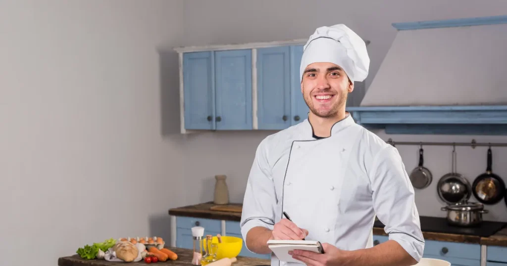 Chef in a white, high-quality uniform and hat, standing in a Sharjah restaurant kitchen with professional cooking tools.