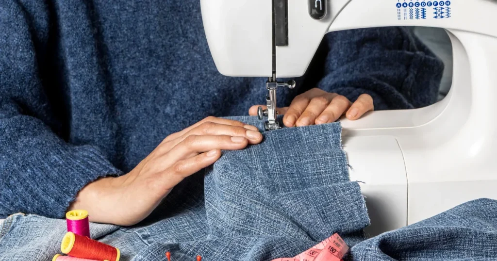 Close-up of hands guiding denim fabric through a sewing machine, surrounded by colourful spools of thread, pins, and a measuring tape, showcasing precise uniform stitching.