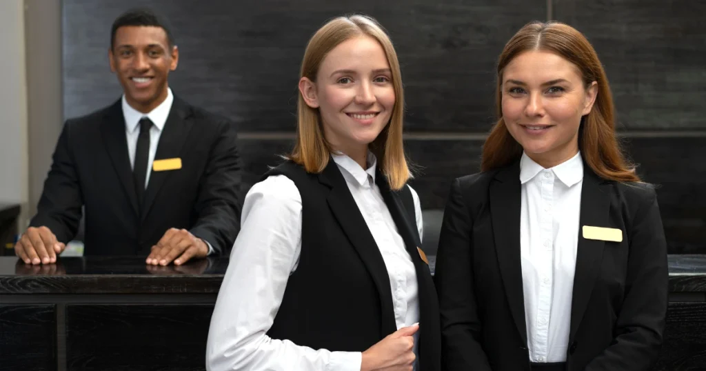 Three hotel reception staff members wearing professional Hotel Industry Uniforms, including black blazers and white shirts, standing behind the reception desk in a modern hotel lobby.