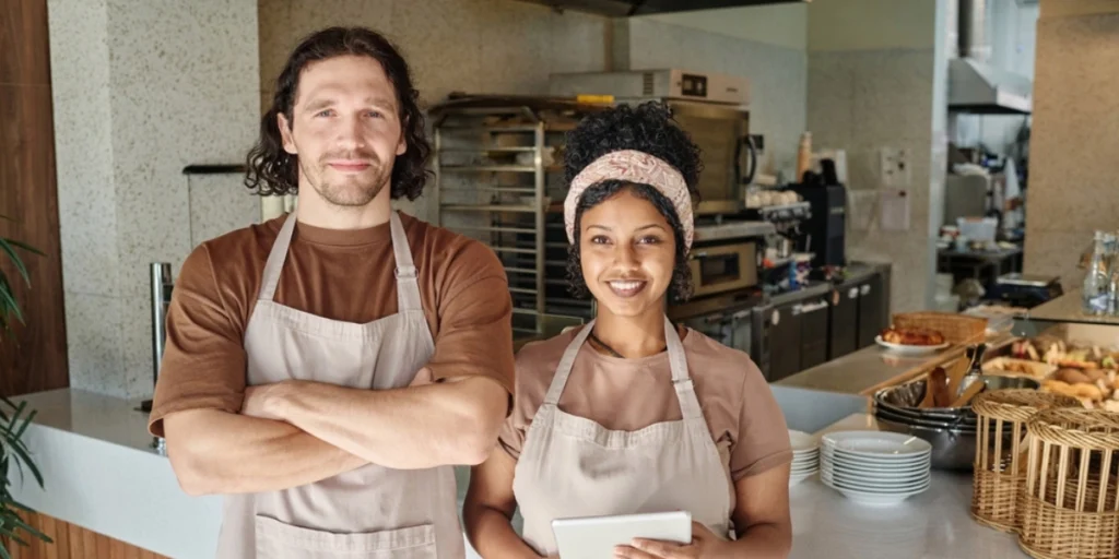 Smiling café staff holding tablet in café staff uniforms inside a bakery café, uniforms showcase, café staff uniforms UAE.