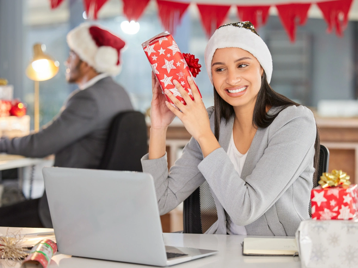 Smiling office worker in Santa hat holding a wrapped present at her desk, celebrating holiday season with christmas corporate gifts uae.